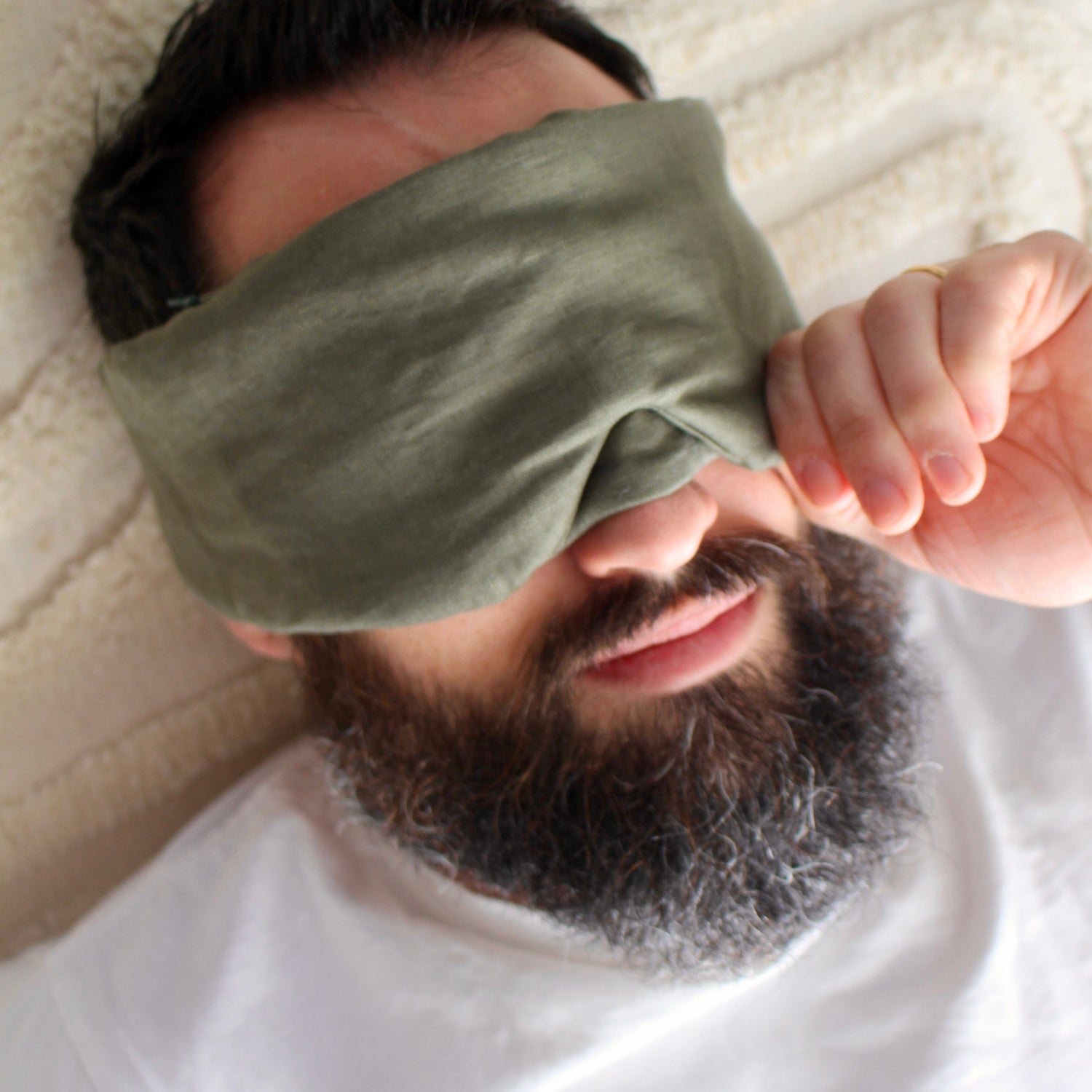 A close-up lifestyle shot of a man lying down with a dark olive green fabric sleep weighted aromatherapy blockout eye mask covering his eyes, wearing a white t-shirt and resting on a textured cream blanket.