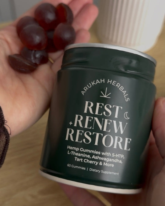 A hand holds a dark green jar labeled 'ARUKAH HERBALS REST RENEW RESTORE' alongside several dark red gummy candies. The background shows a light-colored ribbed mug and a wooden surface.